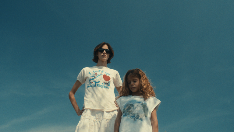 Woman wearing sunglasses and a San Francisco t-shirt stands behind a young girl with curly hair against a blue sky.