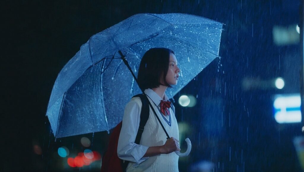 A student in a school uniform stands under a clear umbrella during a rainy night with blue-toned lighting.