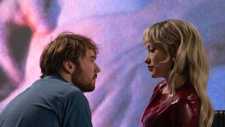 A man in a blue shirt gazes intently at a woman with blonde hair wearing a glossy red top against a blurred purple backdrop.