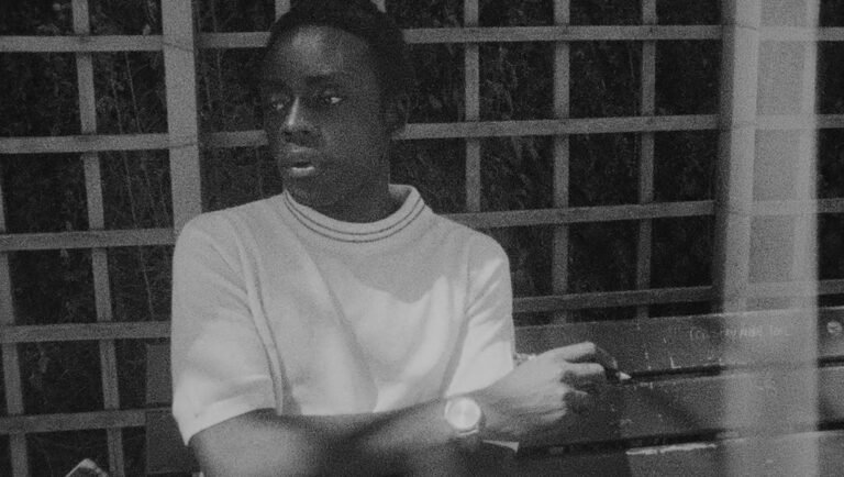 Young man in a white collared shirt sits on a park bench before a wooden lattice, looking away with a thoughtful gaze.