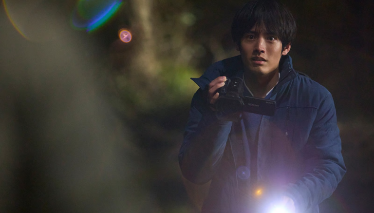 Young man in a blue jacket looks startled while aiming a camcorder and flashlight into the dark woods.