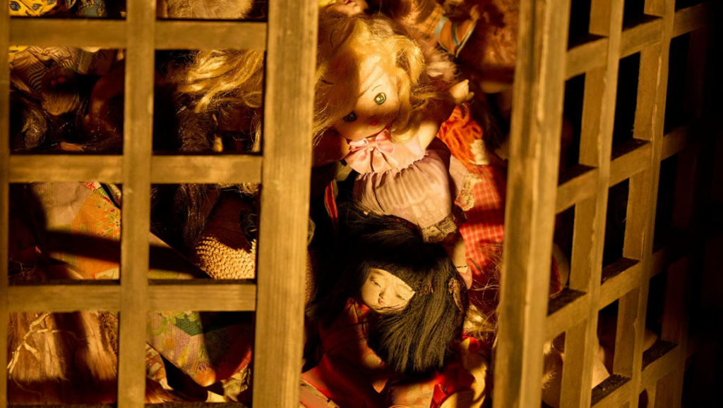 Collection of vintage dolls with varied hair and clothing crowded behind a wooden lattice screen in warm amber light.
