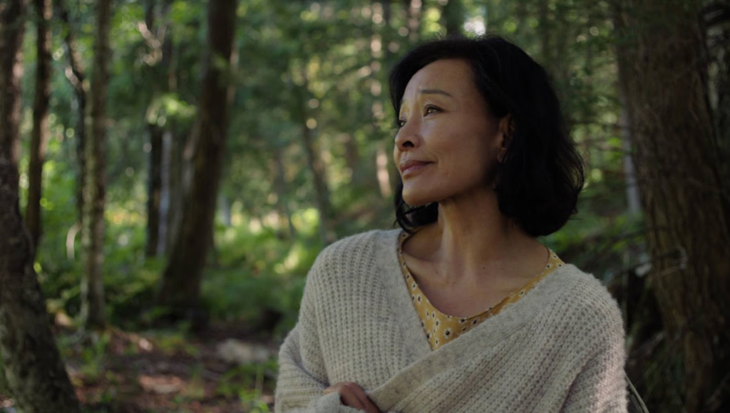 Middle-aged Asian woman in a beige cardigan looking thoughtfully into a sunlit forest.
