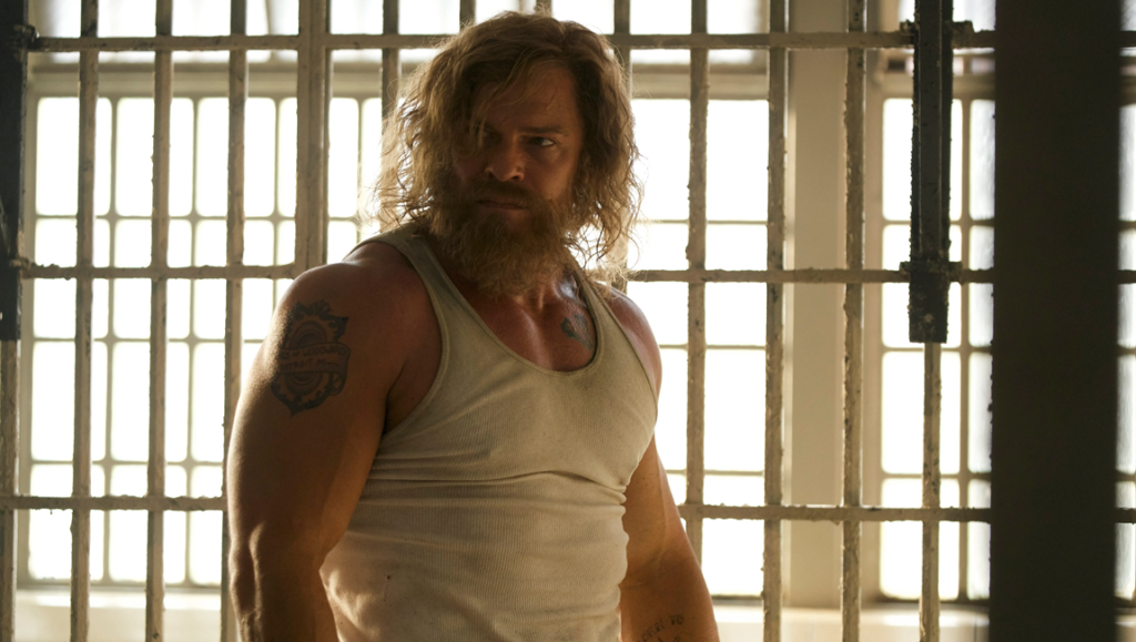 Muscular bearded man with long hair wearing a white tank top stands before a bright, barred prison window.