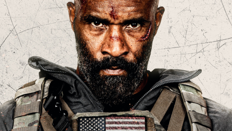 Close-up of a rugged man with a beard and facial wounds wearing a tactical vest with an American flag patch.