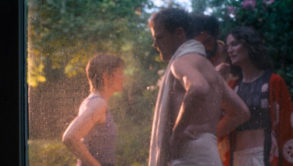 Three people stand outdoors at dusk, seen through a smudged window, while a shirtless man with a towel looks at a woman.