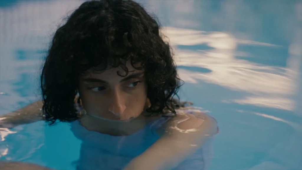 Person with dark curly hair submerged chest-deep in bright blue swimming pool water looks toward the side.