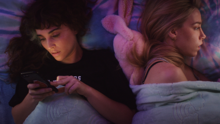 Dark-haired woman scrolls on a smartphone while a blonde woman lies beside her under a blue blanket in purple light.