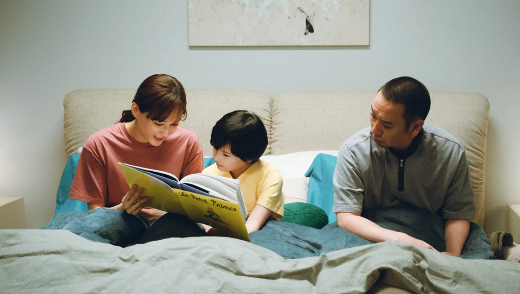 Mother and child reading The Little Prince book together in bed while a father watches.