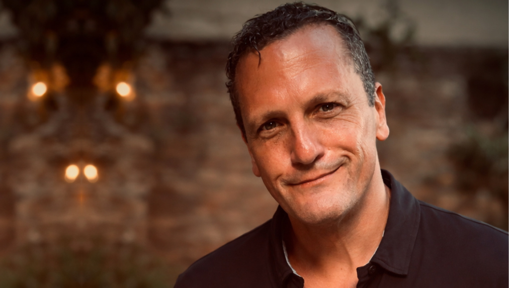 Middle-aged man with short dark hair wearing a black polo shirt, smiling slightly against a blurred warm-toned background.