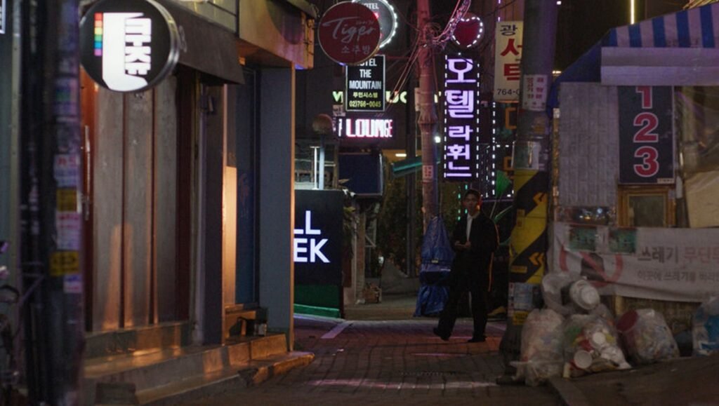 A man walks down a dimly lit, narrow city street at night lined with illuminated Korean neon signs and storefronts.