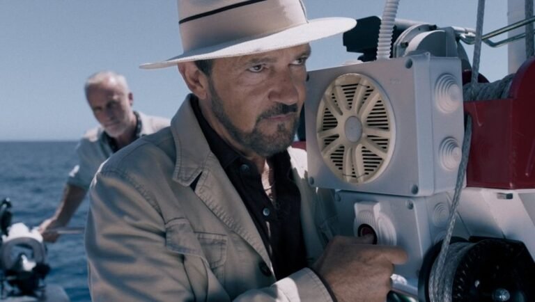 Middle-aged man in a fedora operates equipment on a boat deck with the ocean in the background.