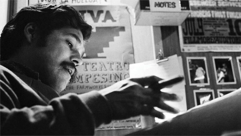 Luis Valdez working at a desk in front of a wall decorated with Teatro Campesino posters in black and white.