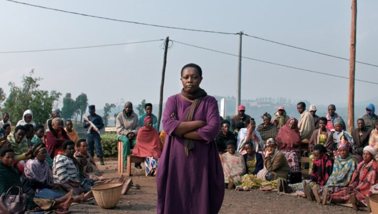 A woman in a purple dress stands with arms crossed before a seated crowd in a dusty, outdoor rural setting.