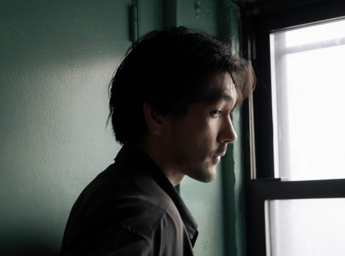 The Negative Version of History: Connor Sen Warnick on Characters Disappearing Connor Sen Warnick stares out a window in profile, illuminated by natural light against a dark green wall.