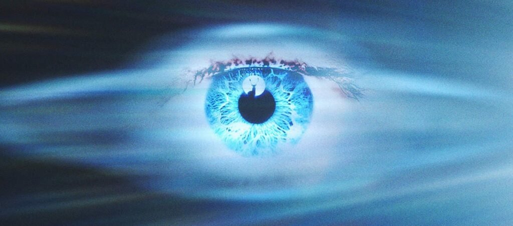 Close-up of a human eye with a bright blue iris reflecting a silhouette of a person standing before a large moon.