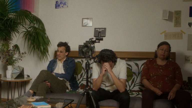Man with headphones covers his face with his hands while sitting on a couch between two other people.