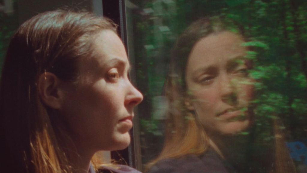 Profile of a woman gazing out a window, her reflection superimposed over blurred green trees passing by.