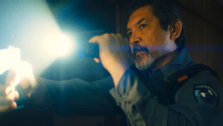 Security guard with a goatee holds a bright flashlight and handgun while investigating a dark, dimly lit environment.