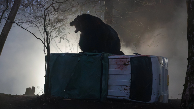 A large dark bear roars while standing atop a white Suzuki truck overturned in a misty, dark forest.