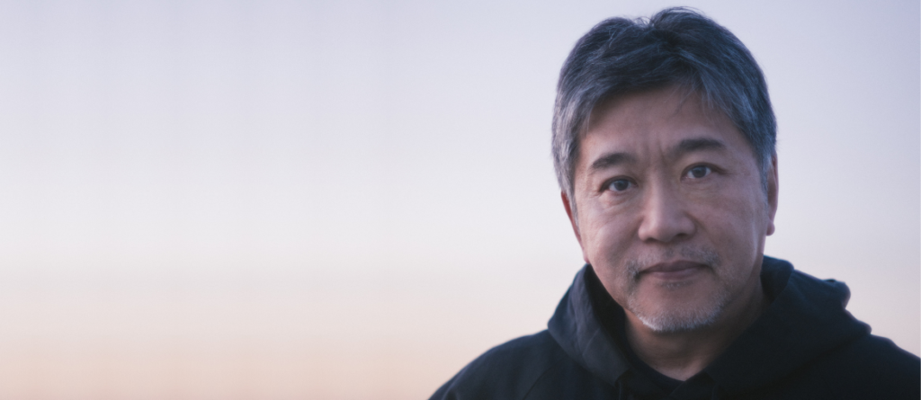 Passing On the Baton: An Interview with Hirokazu Kore-eda on Sheep in the Box Hirokazu Kore-eda with salt-and-pepper hair poses against a soft, pale blue background.