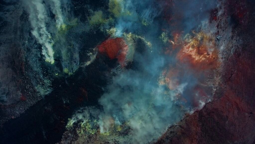 FIDMarseille 2026 — Dispatch 1: Pure Reason, Mirage, Foam of Worlds Aerial view of a volcanic crater releasing thick plumes of smoke over glowing red vents and sulfurous yellow deposits.