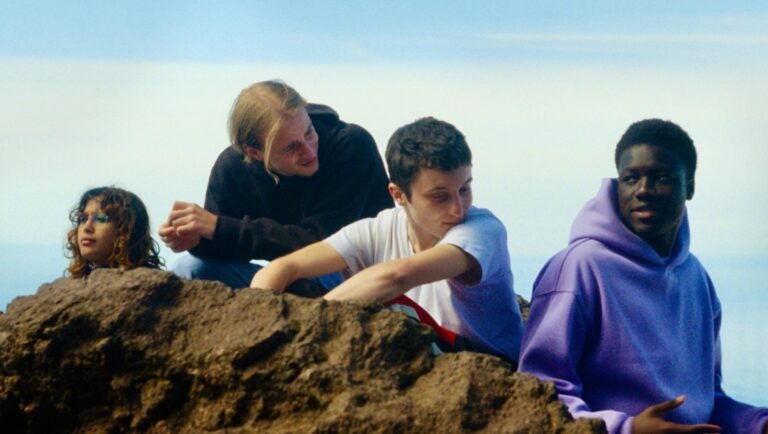 Four diverse young people sit on a rocky outcrop against a bright sky, interacting and looking in different directions.