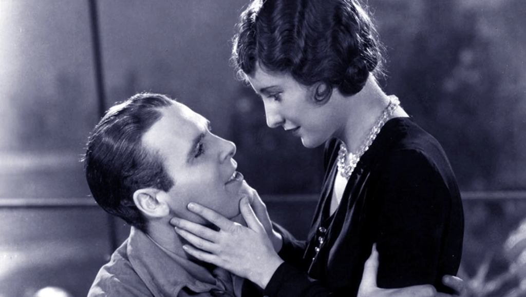 Barbara Stanwyck gazes tenderly at a man while cradling his face in her hands in a black and white film scene.