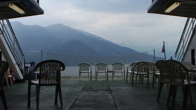 Empty white plastic chairs arranged in rows on a ferry deck overlooking a calm lake and distant misty mountains.