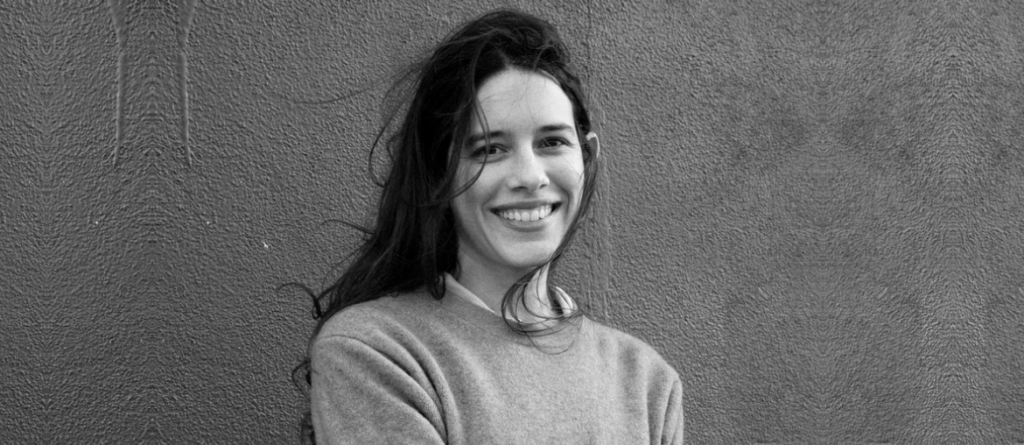 In a Whisper, Not a Scream: An Interview with Lorena Alvarado on Lost Chapters Lorena Alvarado smiling with long dark hair against a textured grey wall.