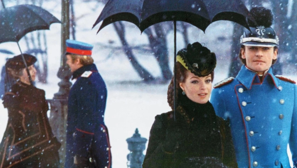 King Ludwig II in a bright blue uniform stands beside a woman in black under falling snow.