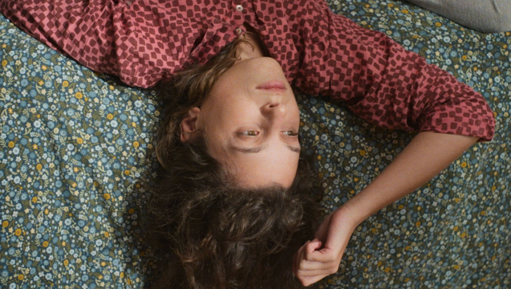 Person with dark curly hair wearing a burgundy checkered shirt lies upside down on a blue floral patterned bedspread.