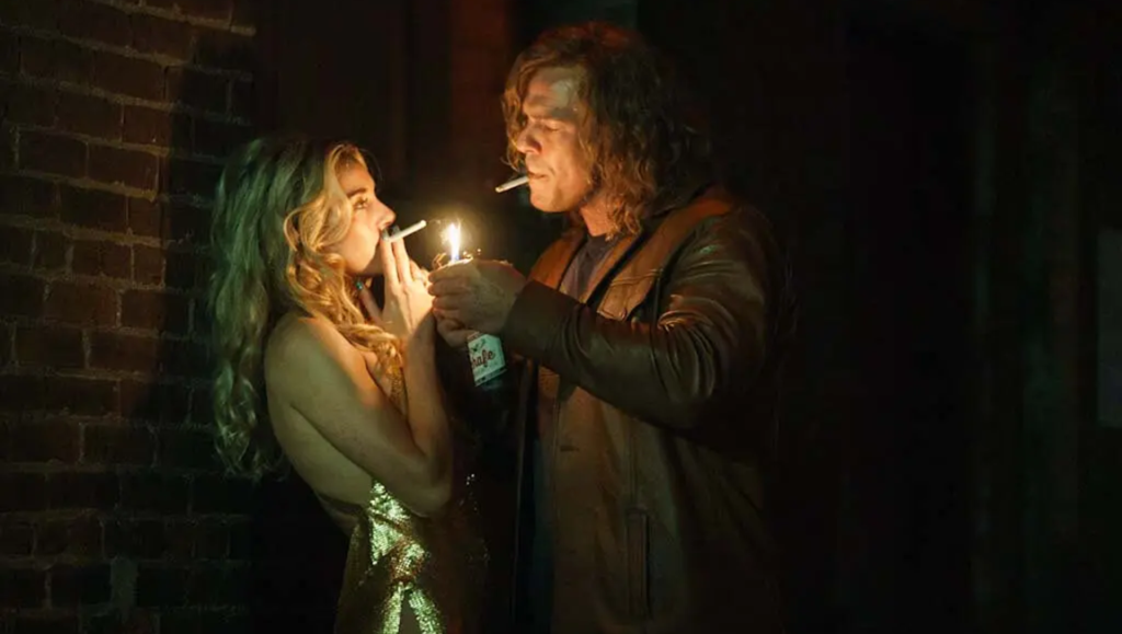 A man in a leather jacket lights a cigarette for a woman in a gold dress against a dark brick wall.