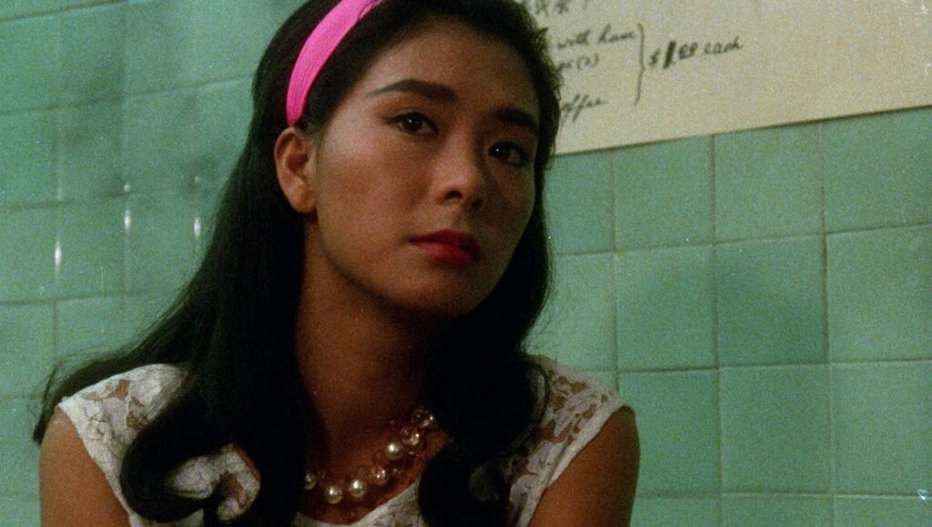 Woman with dark hair, a pink headband, and a pearl necklace sits in front of a light green tiled wall.