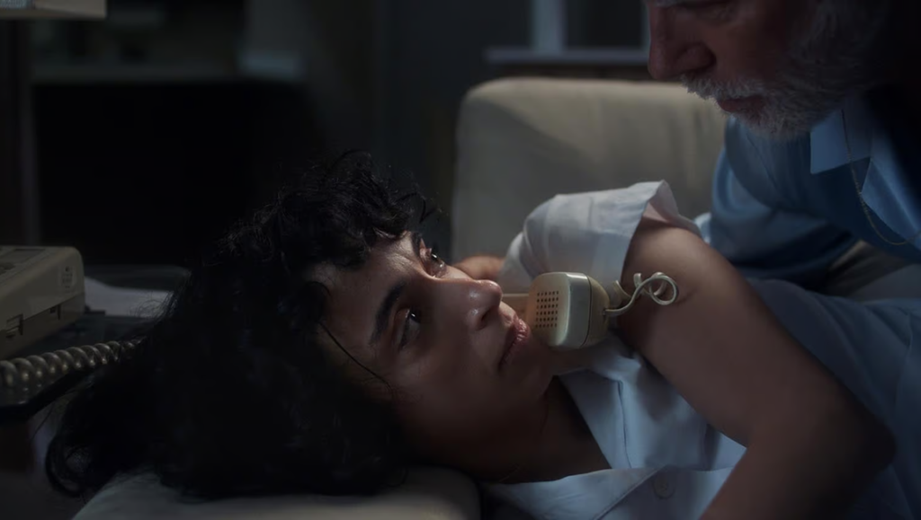 A nurse with curly hair lies on a bed, holding a vintage telephone receiver to her ear while an older man leans over.