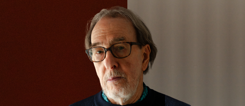 Ross McElwee with graying hair and glasses looking directly at the camera against a brown and gray background.