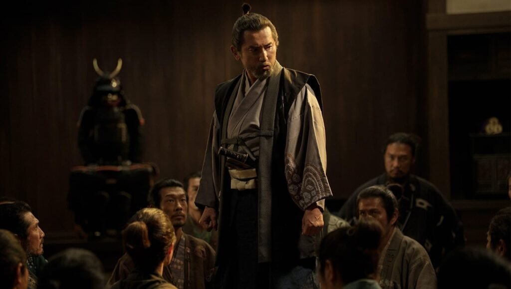 A samurai with a topknot stands sternly before a dimly lit room of seated men with a suit of armor in the background.