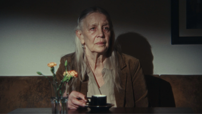 Elderly woman with long grey hair holds a small black cup while sitting at a table with orange carnations.