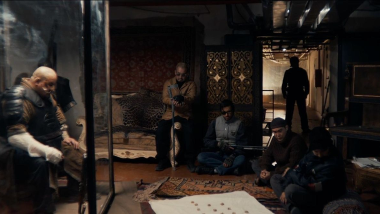 Five men gather in a dimly lit, patterned room to play a board game on a white sheet laid across a rug.