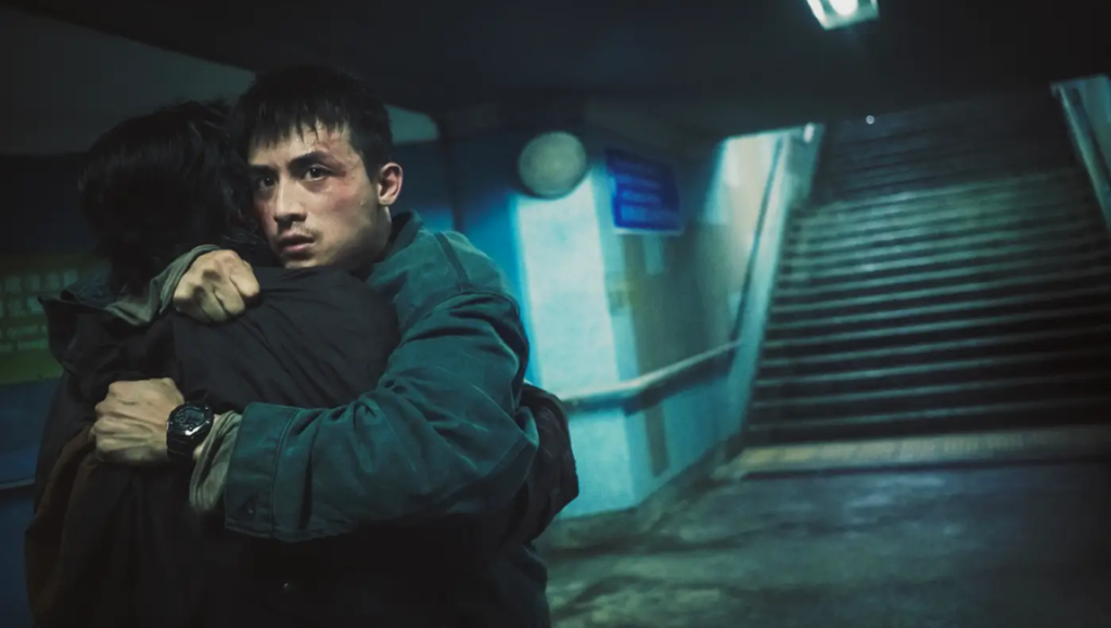 A man with a worried expression hugs another person in a dimly lit, blue-toned subway underpass.