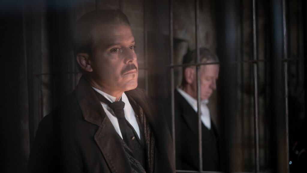 Serious man with a mustache in a dark coat stands behind iron prison bars while an older man looks down in the background.