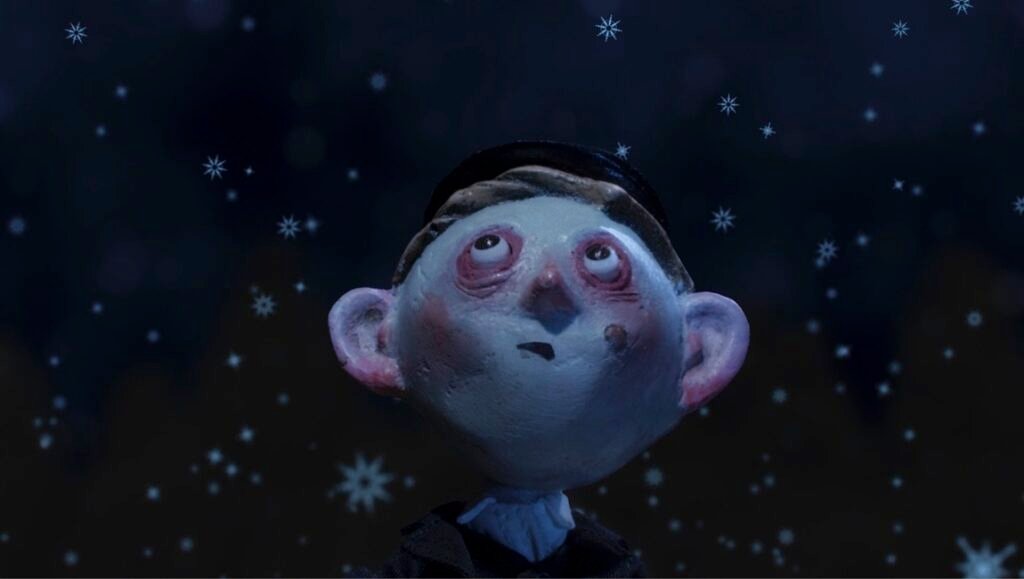 Stop-motion puppet of a boy with pale skin and large ears looking up at falling snowflakes against a dark background.