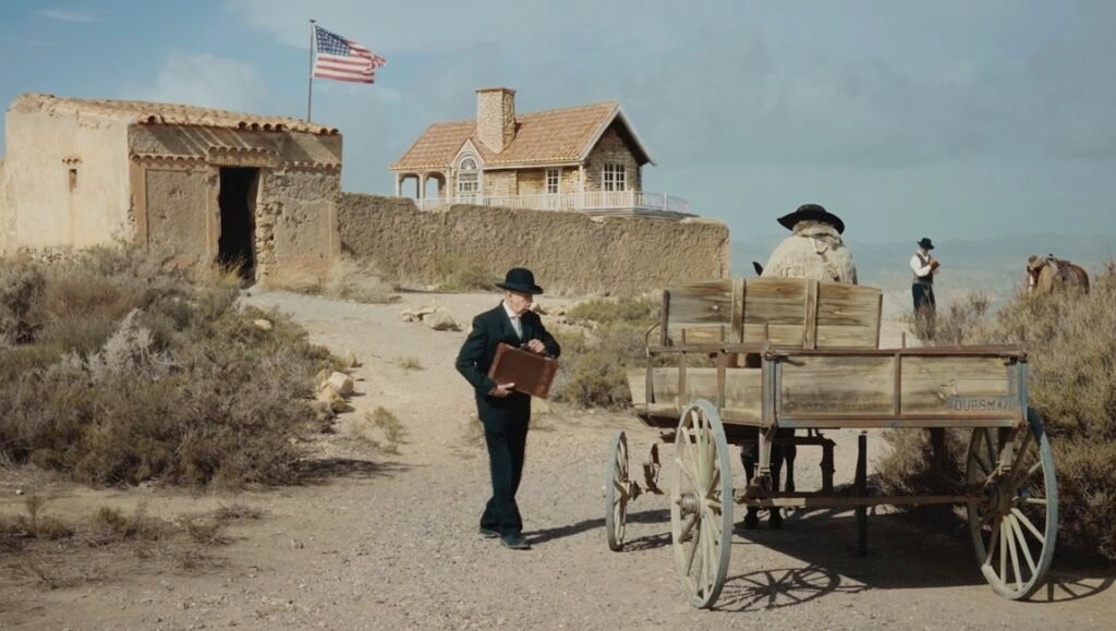 Man in a black suit and bowler hat carries a briefcase past a wooden wagon near a desert ranch house with an American flag.