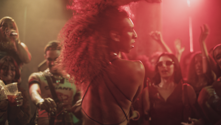 Curly-haired individual dances with their back to the camera amidst a crowded, dimly lit club bathed in red light.