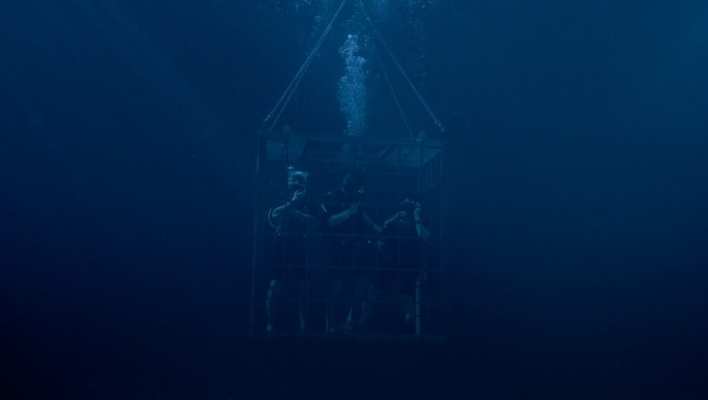 Three scuba divers stand inside a submerged metal cage as air bubbles rise through the deep blue ocean water.