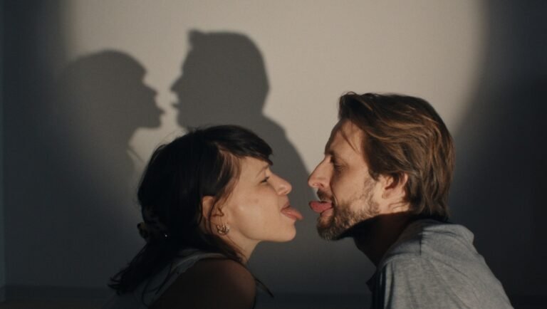 A woman and a man face each other and stick out their tongues, casting shadows on the wall behind them.