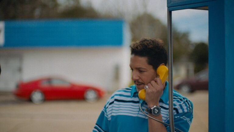 Man wearing a striped blue polo shirt talks on a bright yellow payphone outside a building with a blurred red car.