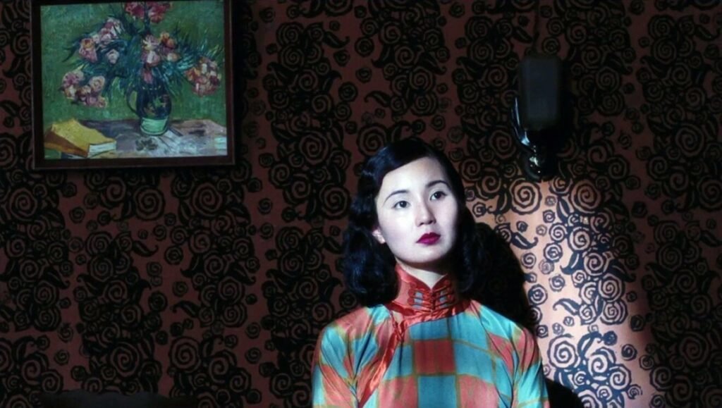 Woman with dark hair and bold lipstick wears a patterned cheongsam against dark floral wallpaper.