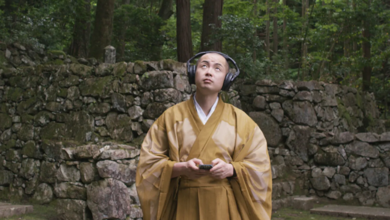 Buddhist monk in golden robes wearing headphones looks upward while holding a smartphone in a forest setting.