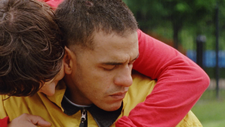 Young man in a yellow jacket with his eyes closed being hugged from behind by someone wearing a red long-sleeve shirt.